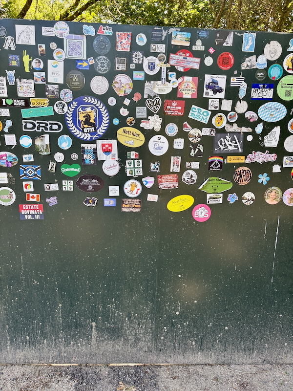 Dark green wall densely covered with colorful stickers and logos outdoors, with trees above.