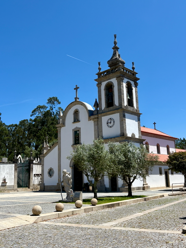 Front view of a white church with a bell tower and clock, set in a sunny cobblestone plaza with trees nearby