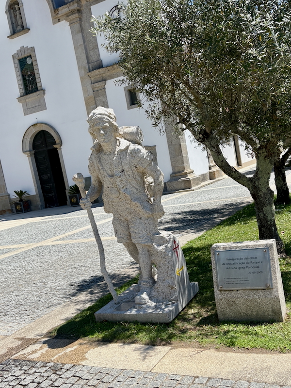 Stone statue of an elderly man with a walking staff standing in a cobblestone plaza beside a white church, with a tree and a plaque nearby.