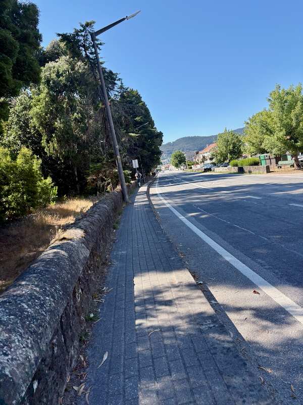 Sunlit sidewalk beside a two-lane road, leaning streetlight, and green trees with hills in the distance.