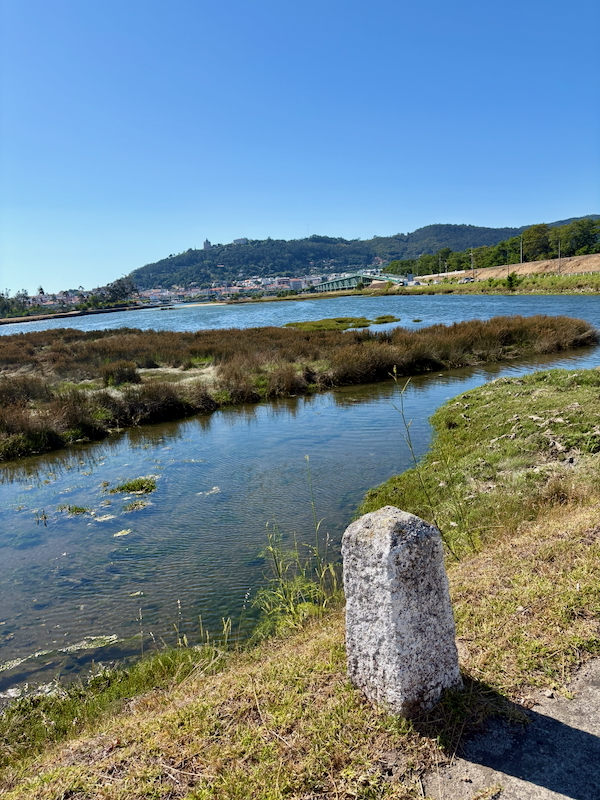 Riverside scene with marshy inlets, a stone bollard in the foreground, a bridge in the distance, and hills under a clear blue sky.