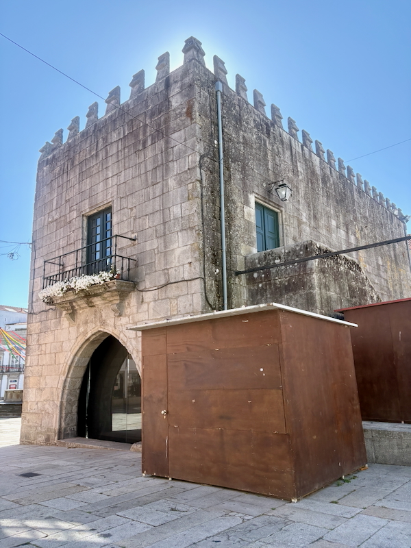 Historic stone building with a crenellated roofline, arched entrance, and small balcony with flowers on a sunny plaza.
