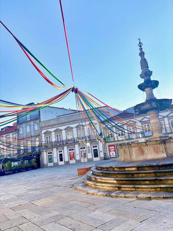 Square with a tiered stone fountain on the right and colorful ribbons radiating from a central point above the plaza.