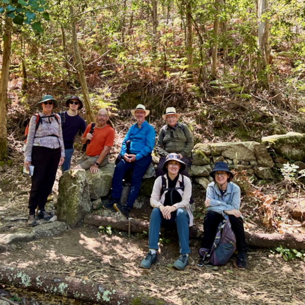 Group of seven hikers resting on large rocks along a forest trail, wearing hats and backpacks on a sunny day.