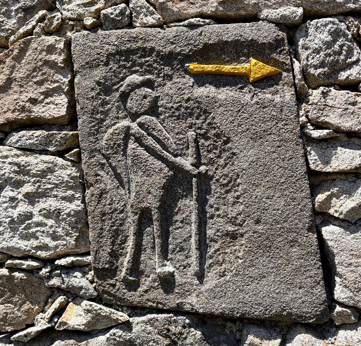 Stone wall with a carved figure resembling a person and a small yellow arrow pointing to the right on the rock surface.