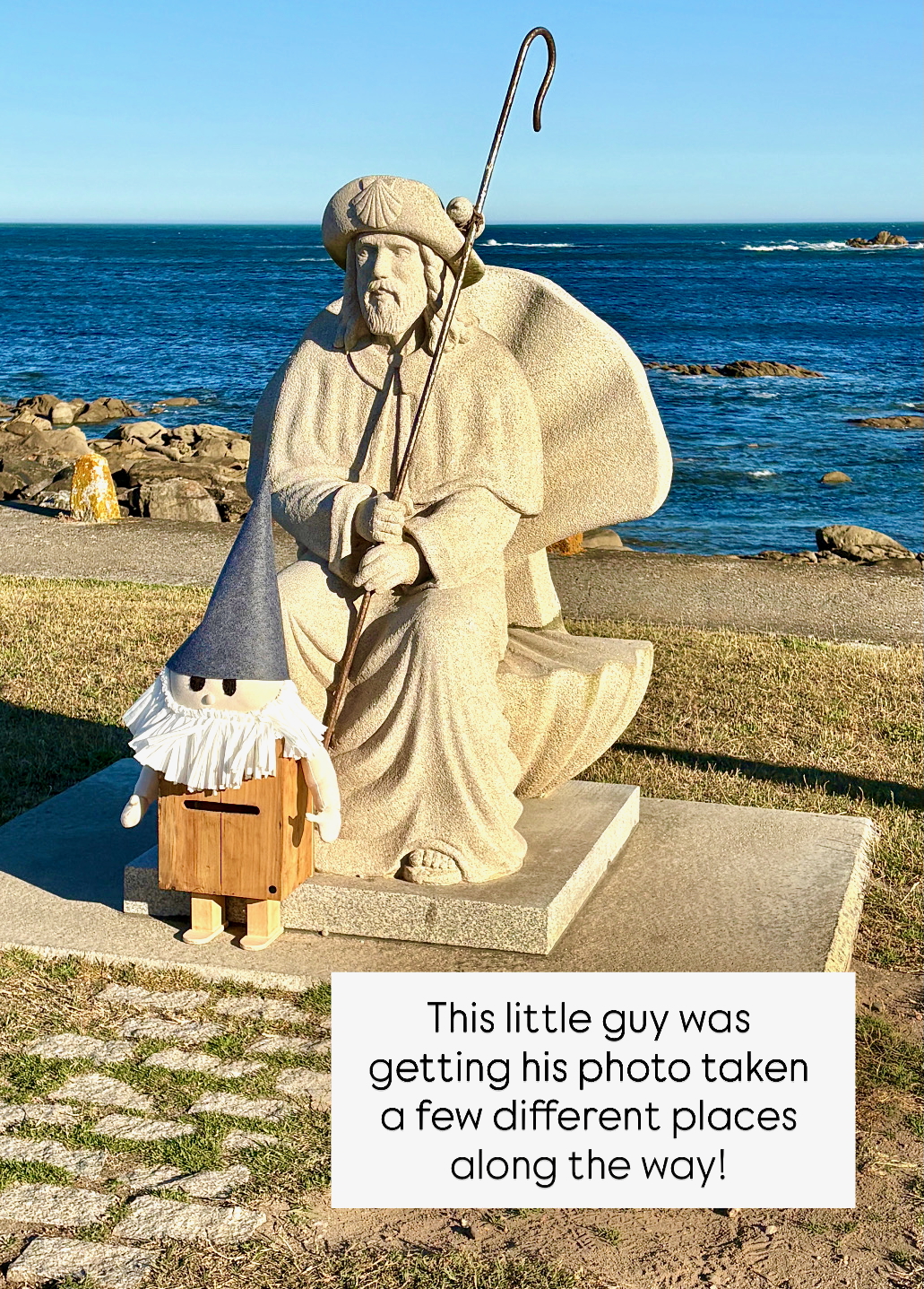 Stone statue of a bearded man in robes with a walking staff by the rocky seaside, accompanied by a small wooden gnome figure.