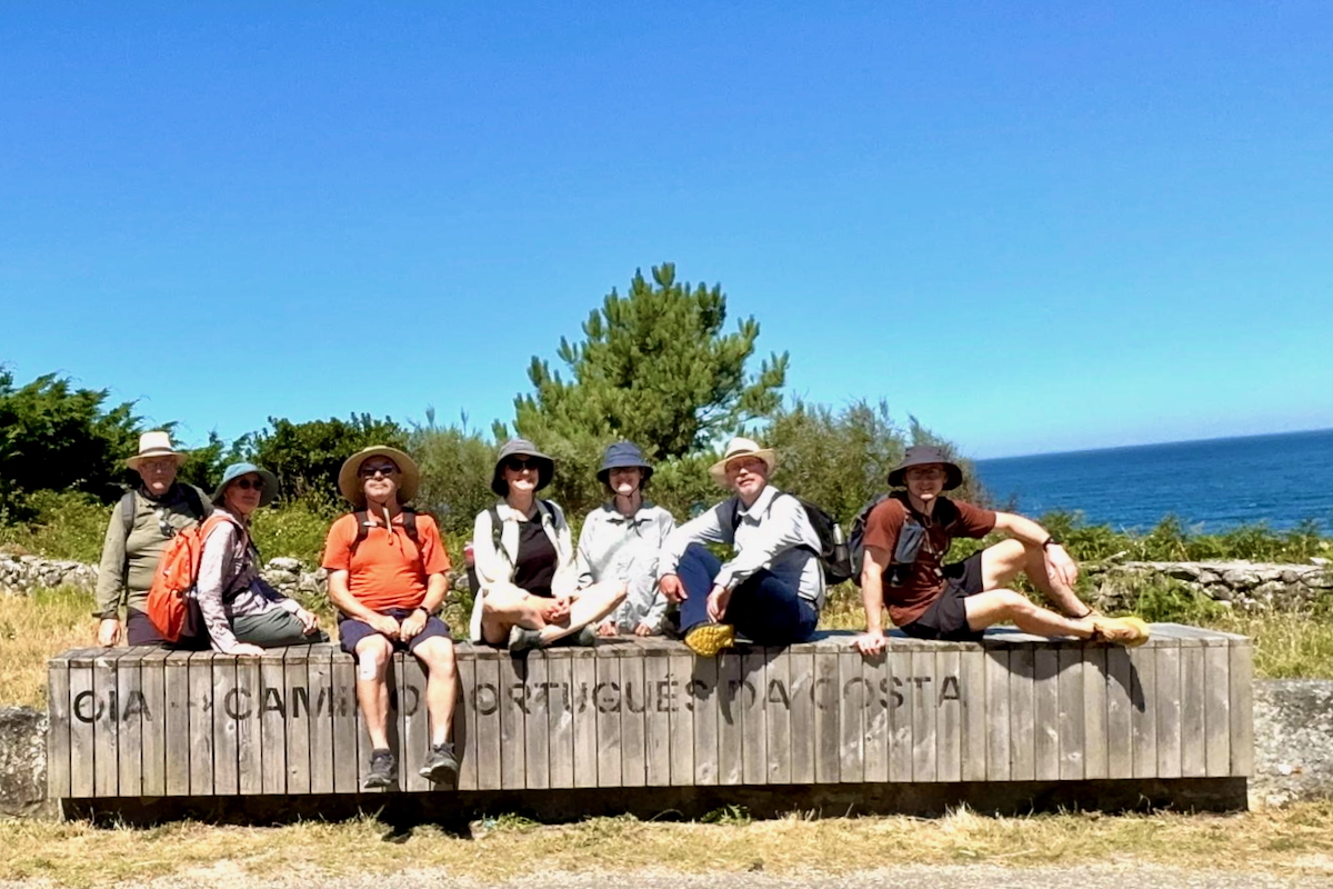 Seven hikers sit on a weathered wooden bench by the coast, with blue sky and ocean behind them.