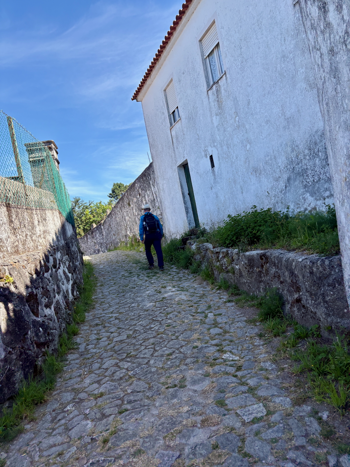 Hiker with backpack walking up a cobblestone street between a white building and a stone wall under a blue sky.