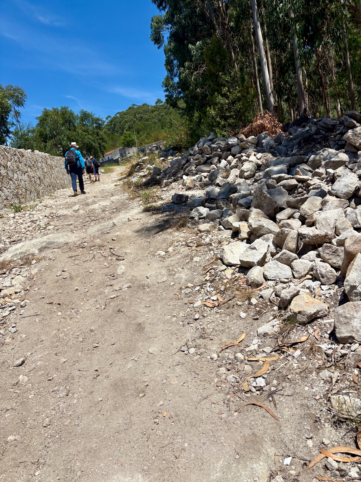 Hikers walking along a dirt rural path beside a stone wall and a pile of rocks under a clear blue sky.