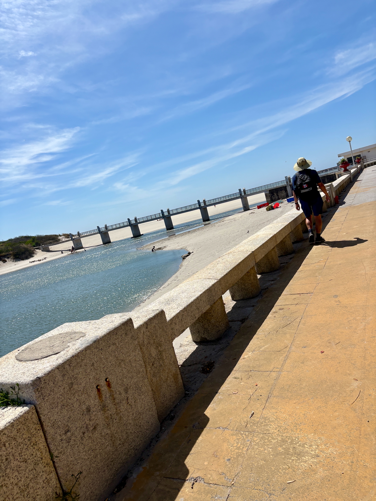 Man wearing a hat and backpack walks along a concrete seawall beside a calm bay on a sunny day