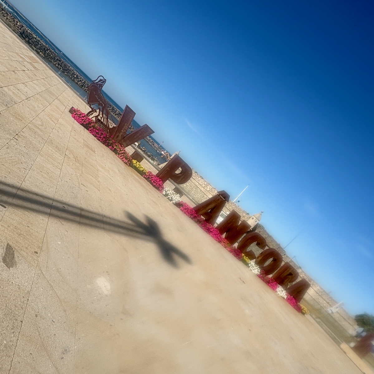 Large rusty letters spell out a place name along a seaside promenade with flower beds and a metal sculpture, under a clear blue sky.