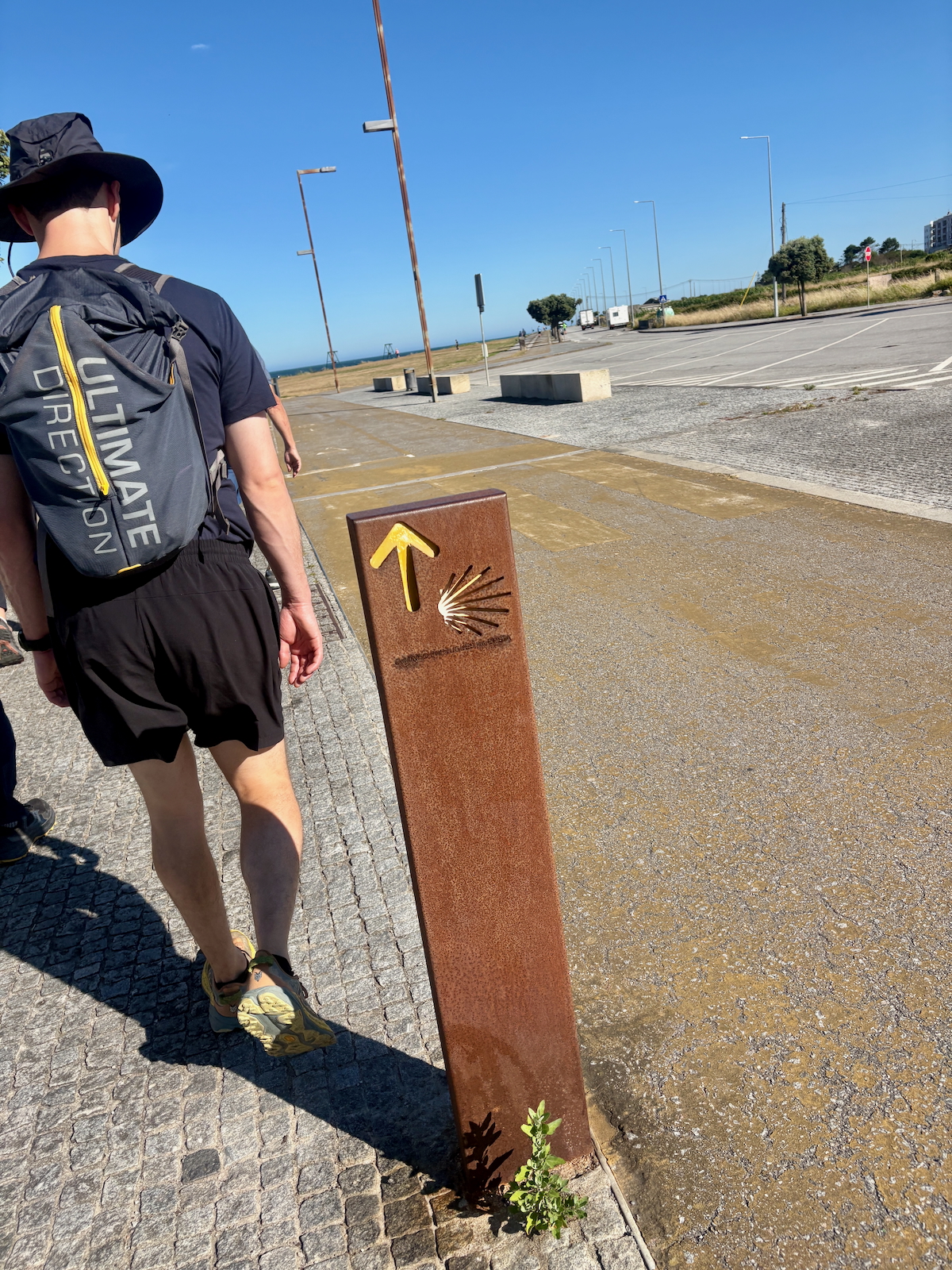 Hiker in a black outfit and hat walks along a coastal promenade; a rusted trail marker with a yellow arrow is in the foreground.