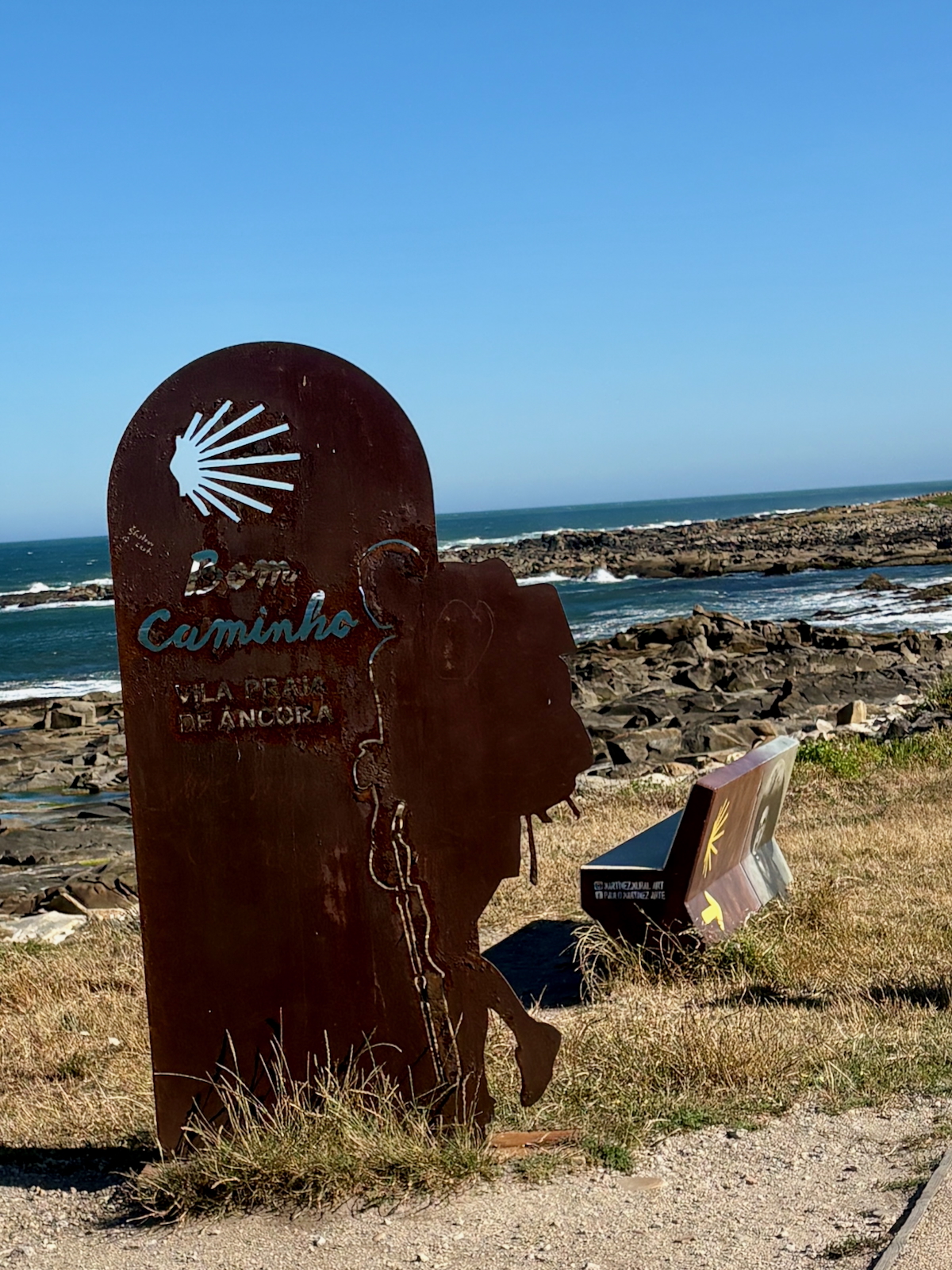 Rusty metal coastal sculpture with a shell motif and the inscription 'Caminho' by a rocky shoreline and blue sea under a clear sky.