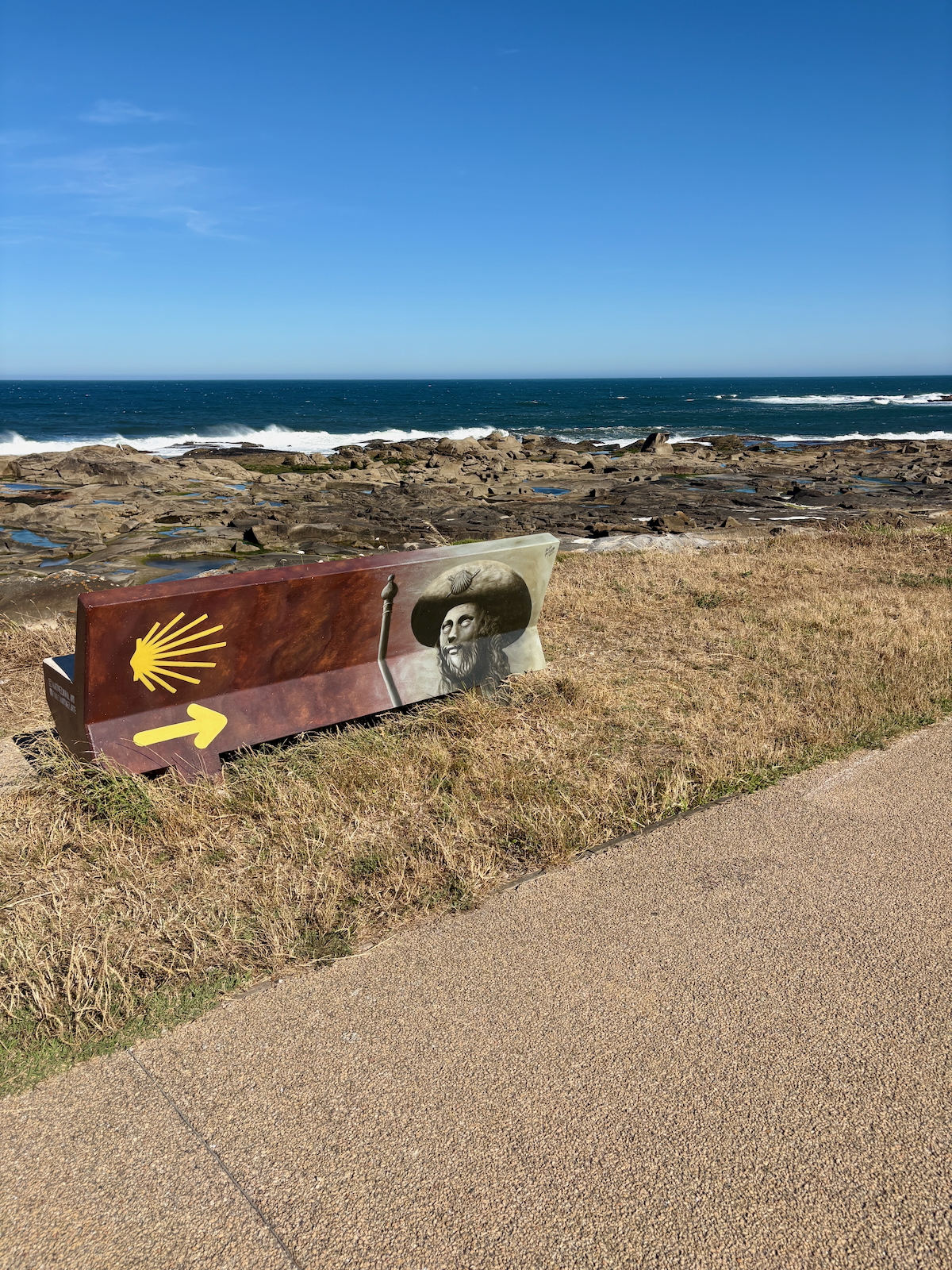 Sign along a rocky coast displays a yellow scallop shell, a left-pointing arrow, and a portrait of a man in a wide-brimmed hat against the ocean backdrop.