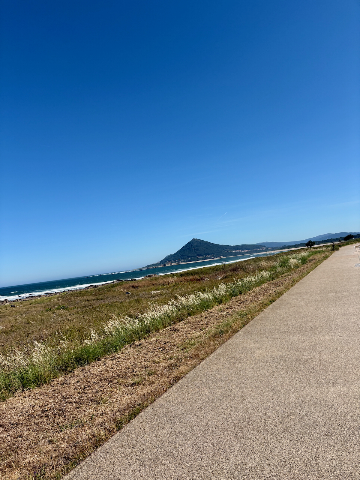 Coastal paved path running along dry grasses, with the blue ocean to the left and a distant hill on the horizon under a clear sky، and distant mountains in the far background.