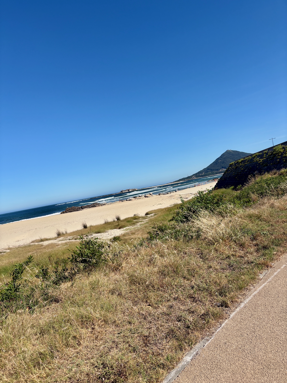 Clear blue sky over a sandy beach with a grassy dune and a sloped hill in the background; a paved path runs along the foreground.