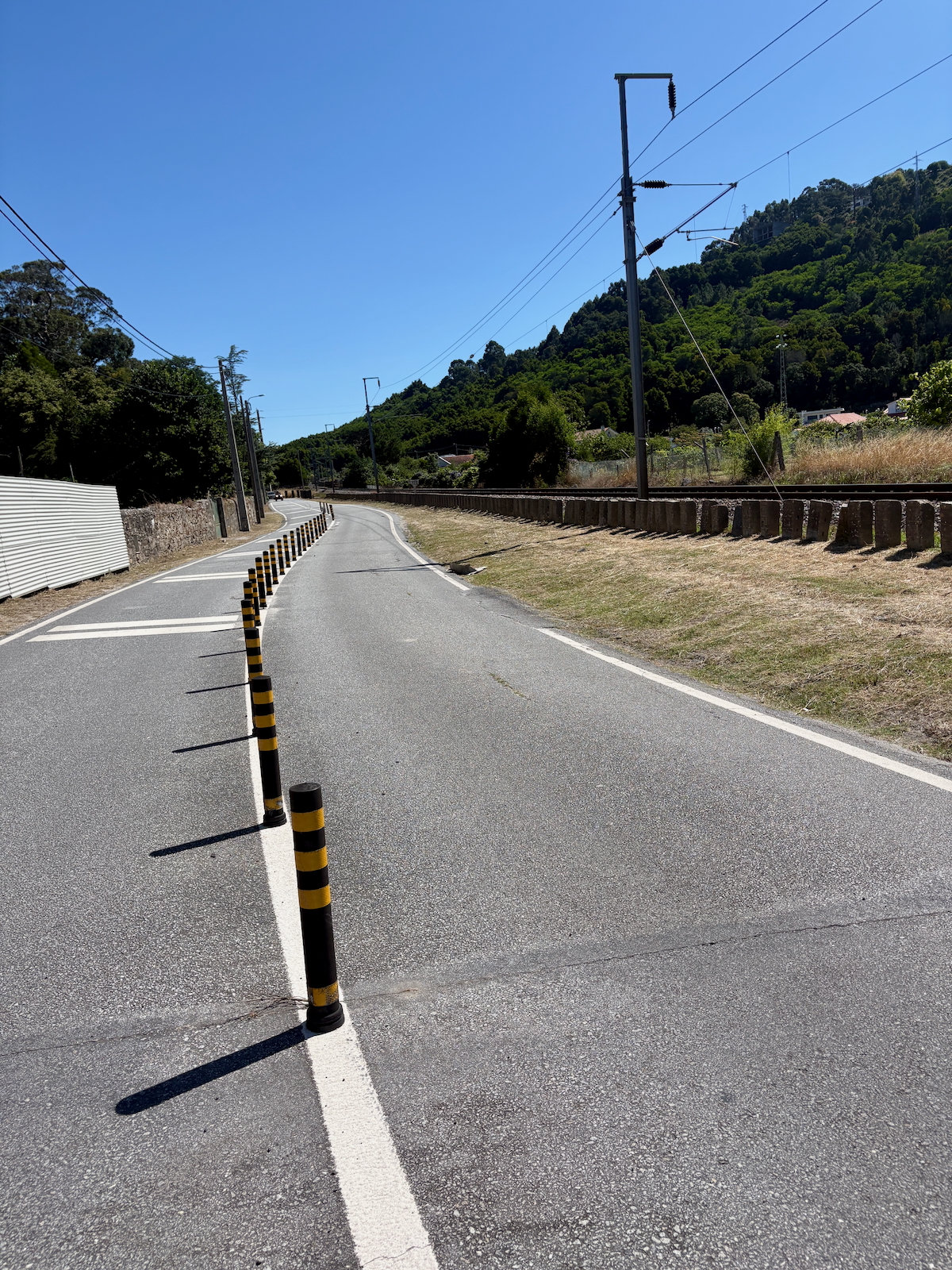 Curving rural road with black-and-yellow bollards along the center and power lines, hillside in the background under a clear blue sky.