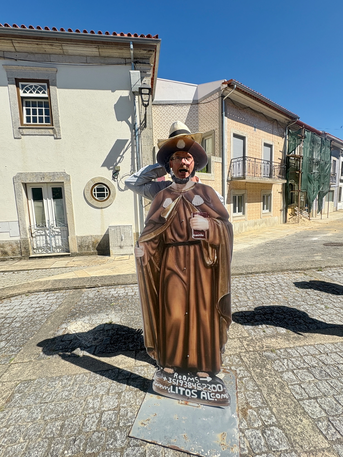 Life‑size cardboard cutout of a monk in brown robes on a pedestal in a sunny cobblestone street with pastel houses behind it.