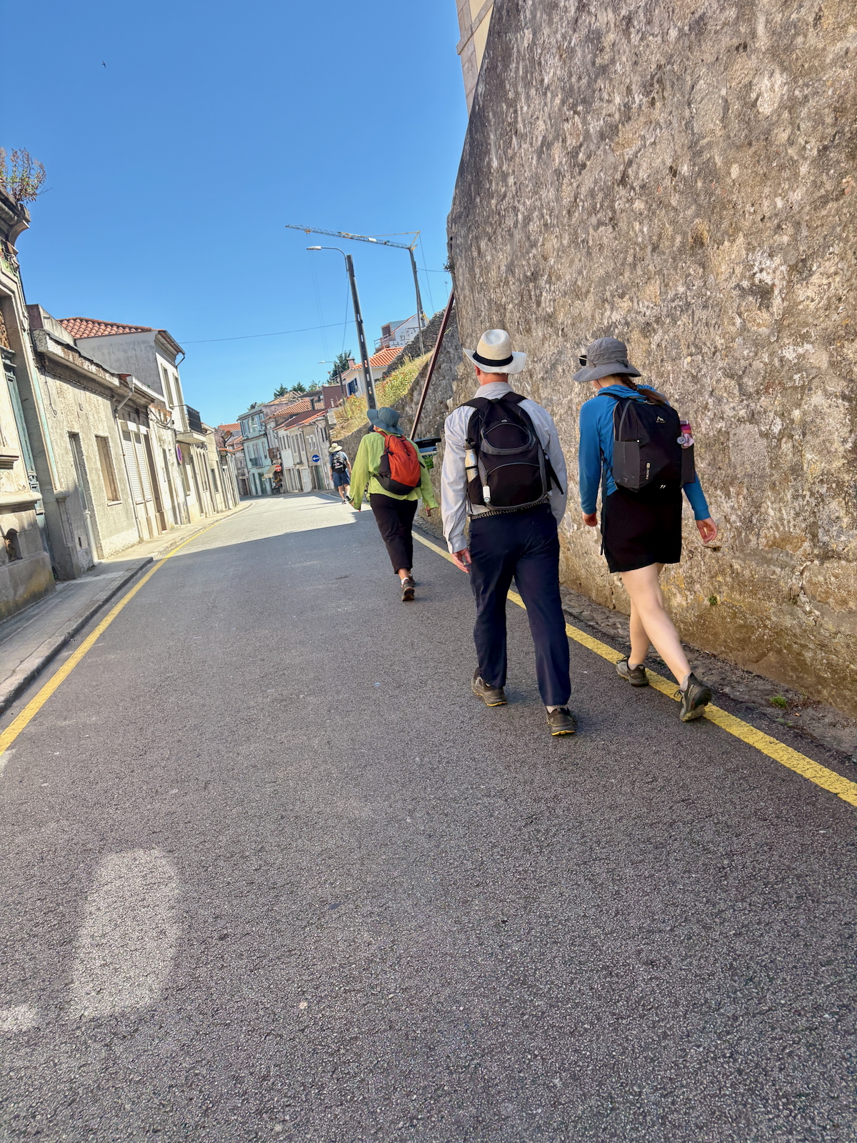 Group of hikers walking away down a sunlit narrow European street, with backpacks and hats, beside an old stone wall and row of houses.