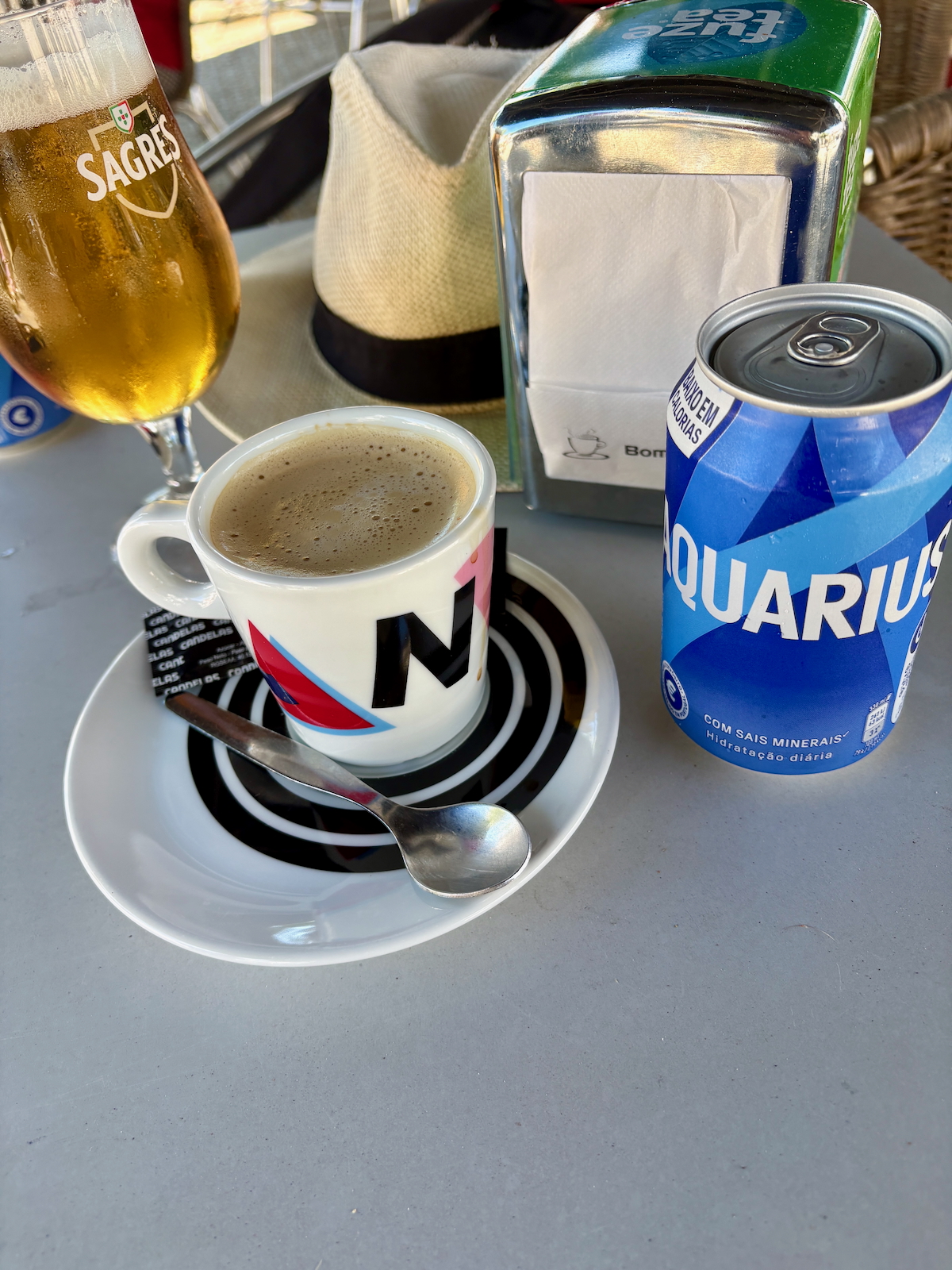 Espresso in a white cup on a saucer with a spoon, beside a blue Aquarius can on a table.