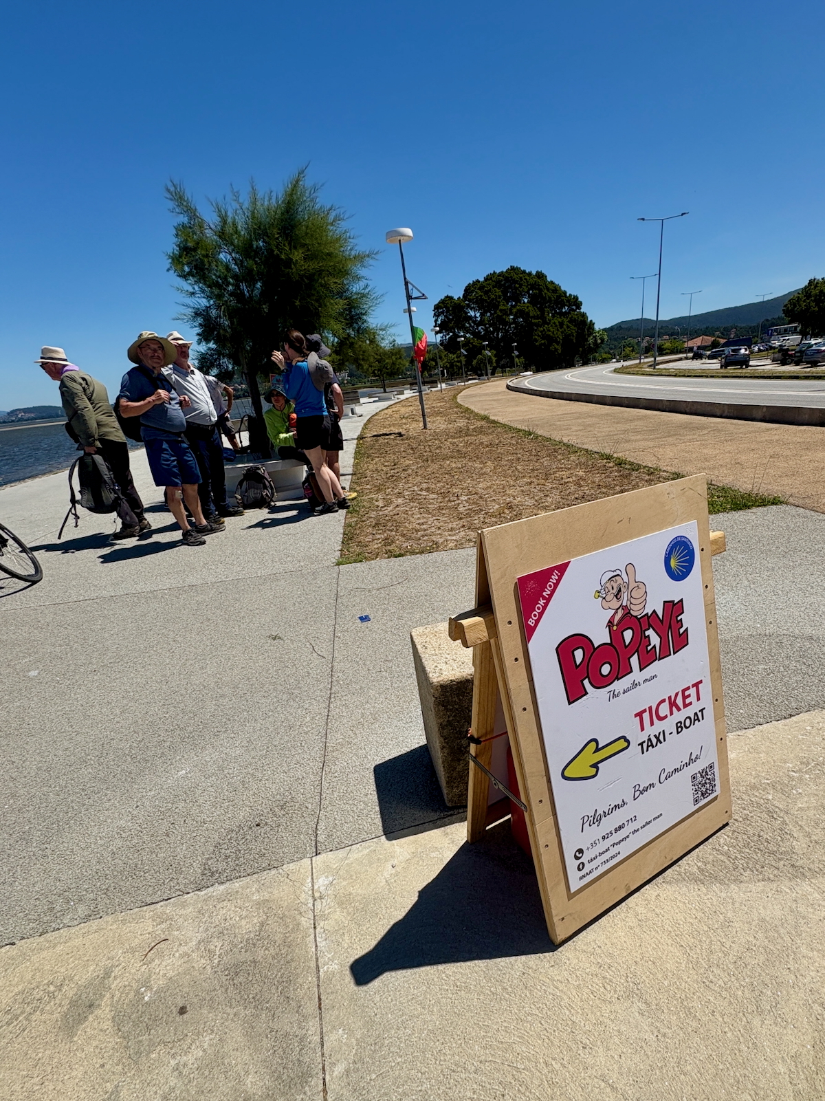 Group of people with backpacks and hats gathered by a riverside promenade as a sign advertises Popeye taxi-boat tickets nearby.