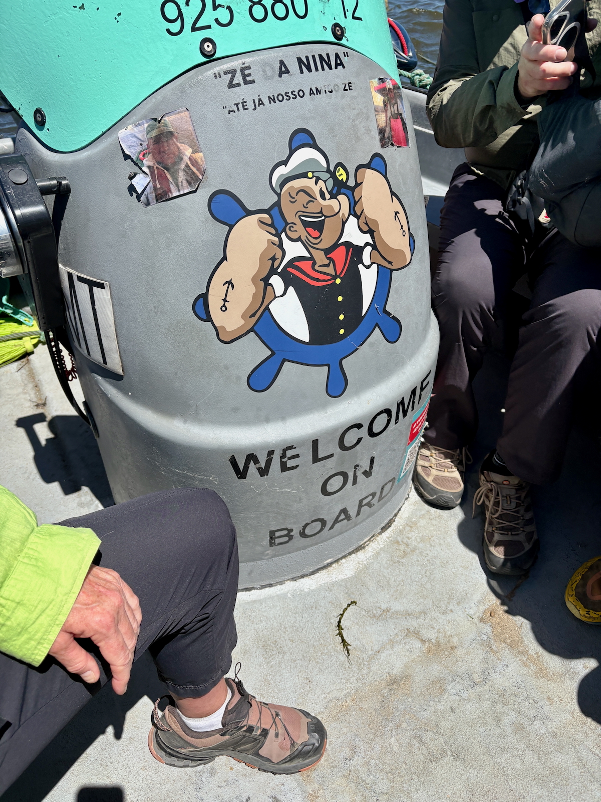 Grey boat hull with large cartoon sailor sticker and 'WELCOME ON BOARD' text, people seated nearby on a sunny deck.