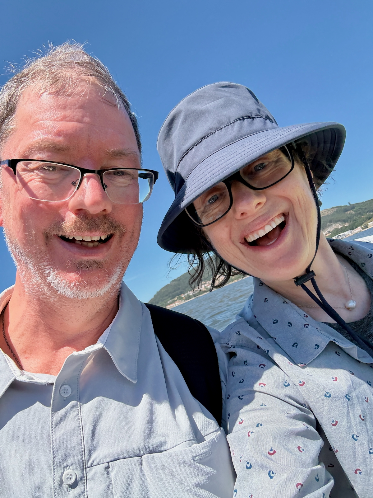 Two friends taking a cheerful selfie outdoors by a lake, with a clear blue sky and hills in the background.