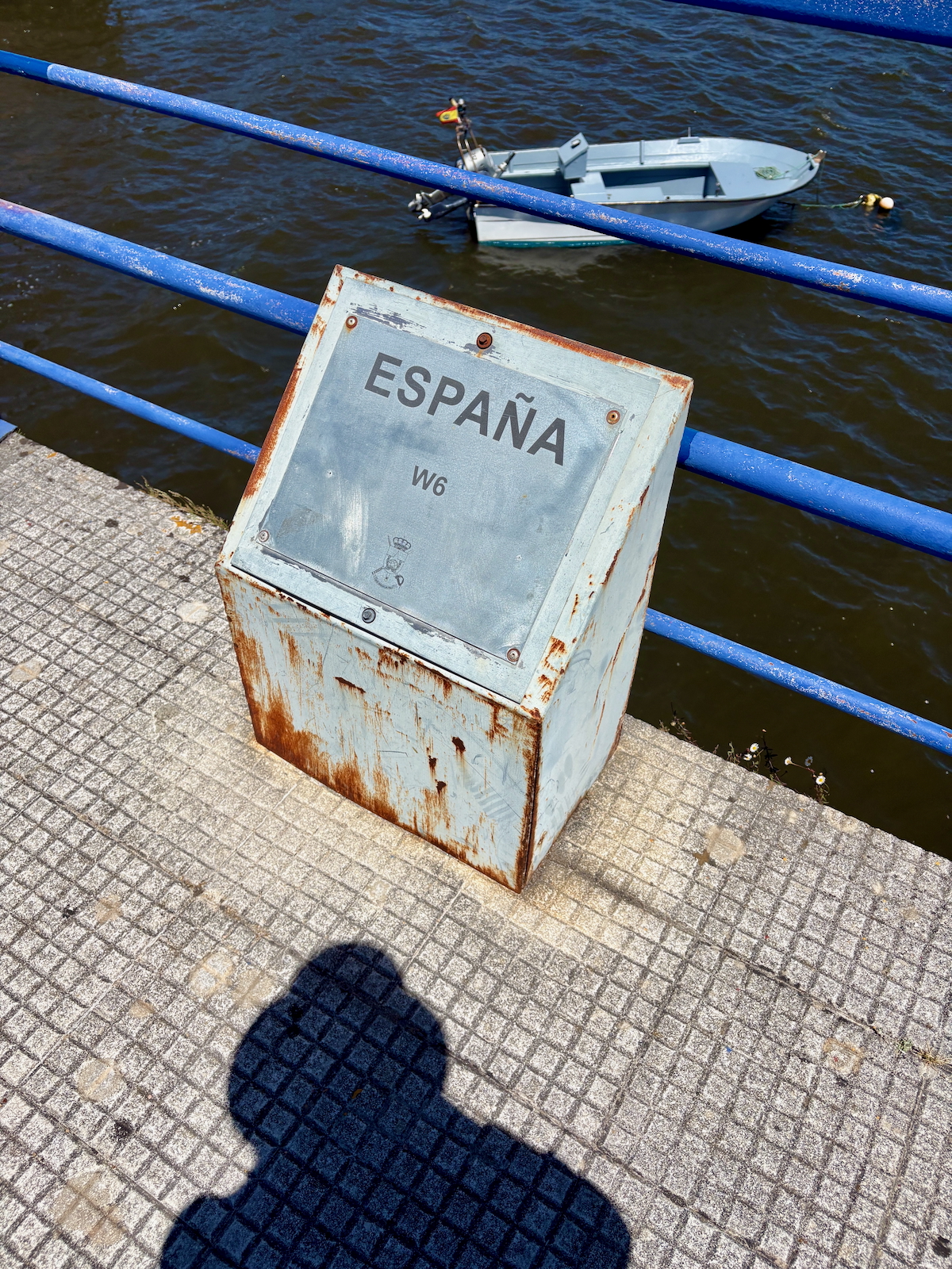 Rusty metal kiosk with the word ESPAÑA and code W6, placed on a concrete walkway beside a blue railing above dark water; a small boat floats in the background.