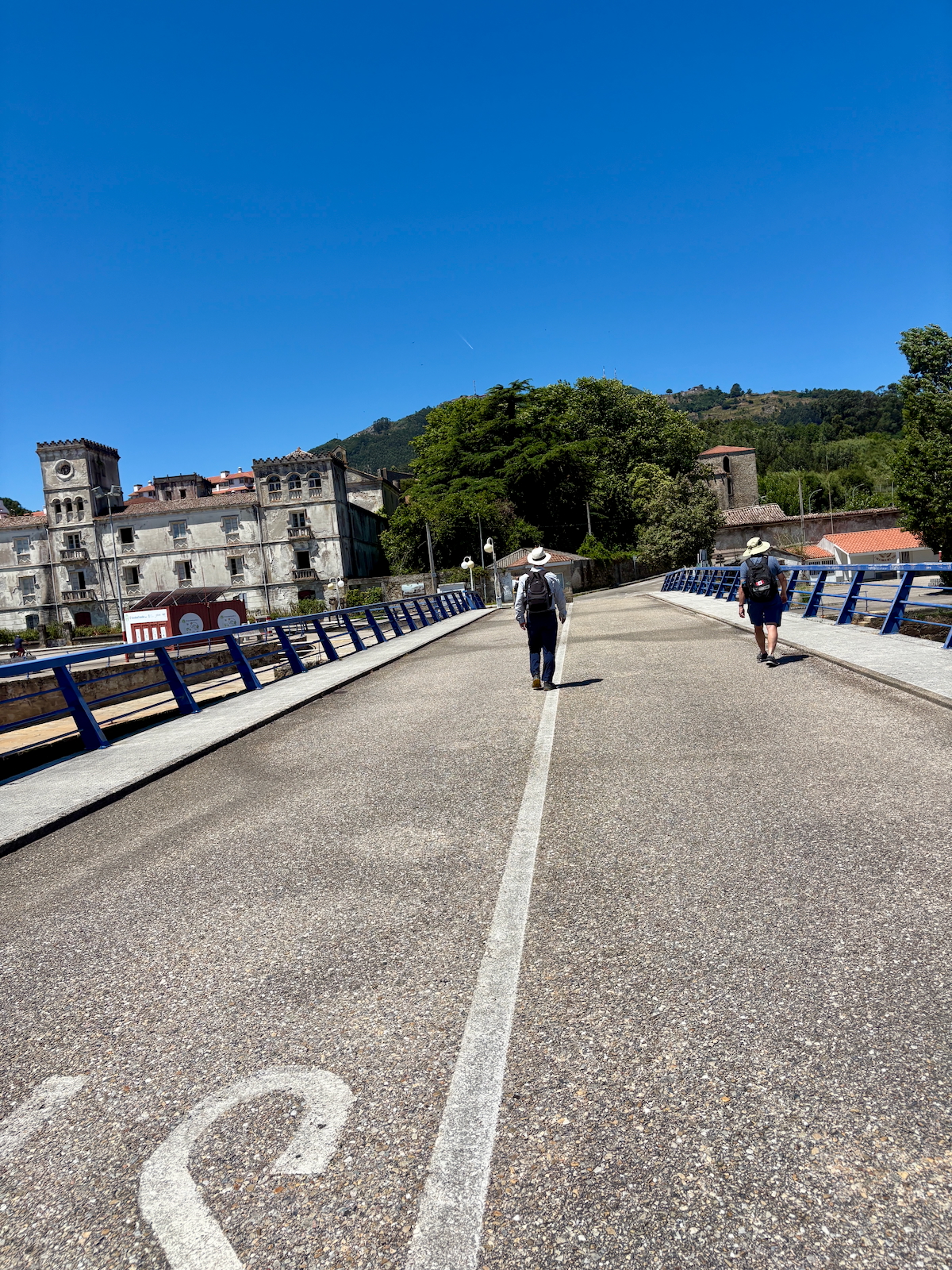 Two hikers with backpacks walk away on a sunny bridge with blue railings; historic buildings line the left side and green hills rise in the distance.