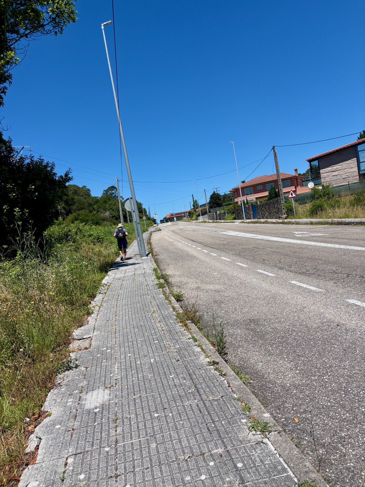 A person with a backpack walks along a cracked sidewalk beside a two-lane road on a sunny day, with houses and streetlights in the background and a bright blue sky.