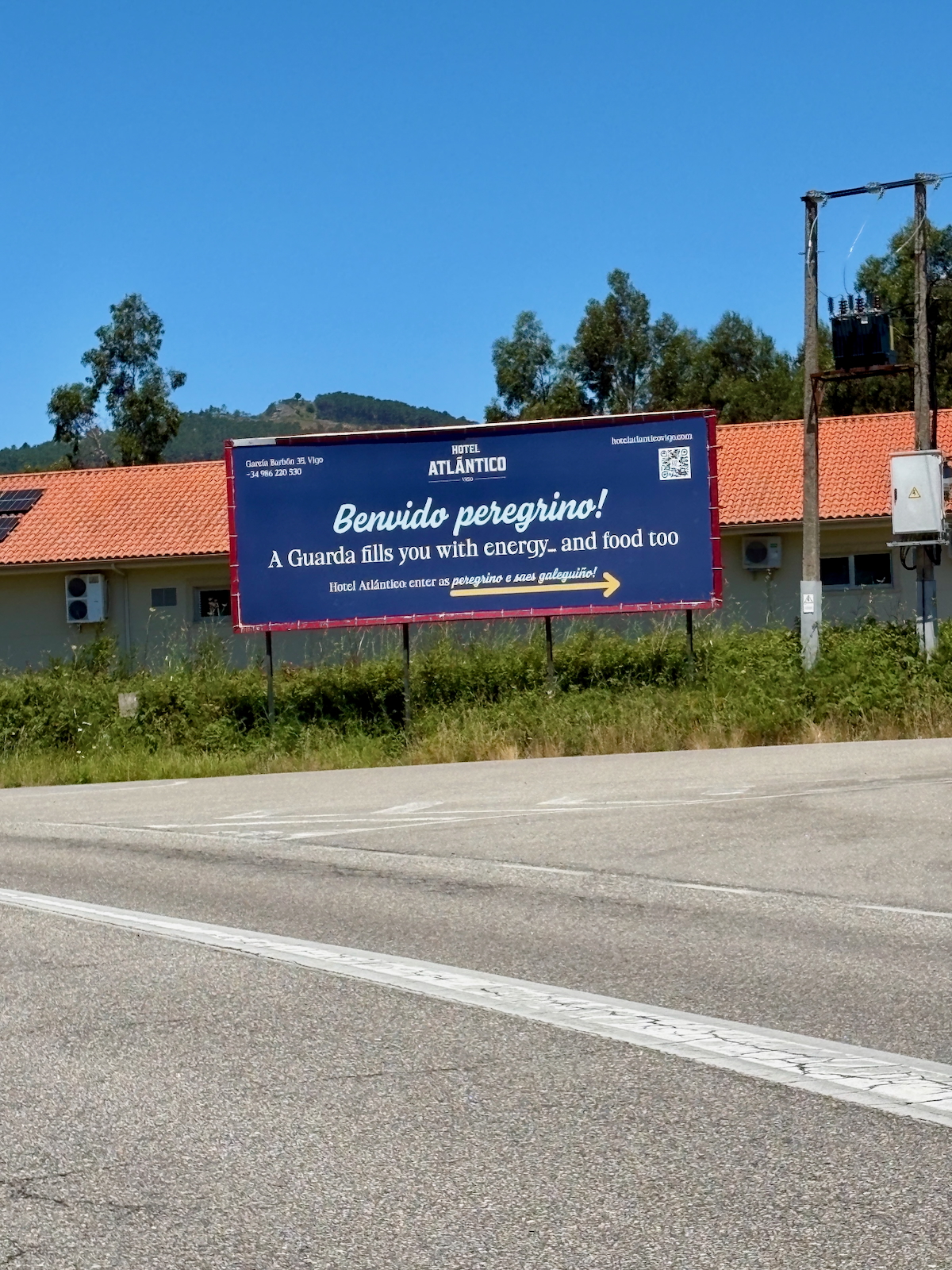 Blue roadside billboard for Hotel Atlántico reading 'Benvido peregrino!' with an arrow, against a building and blue sky.