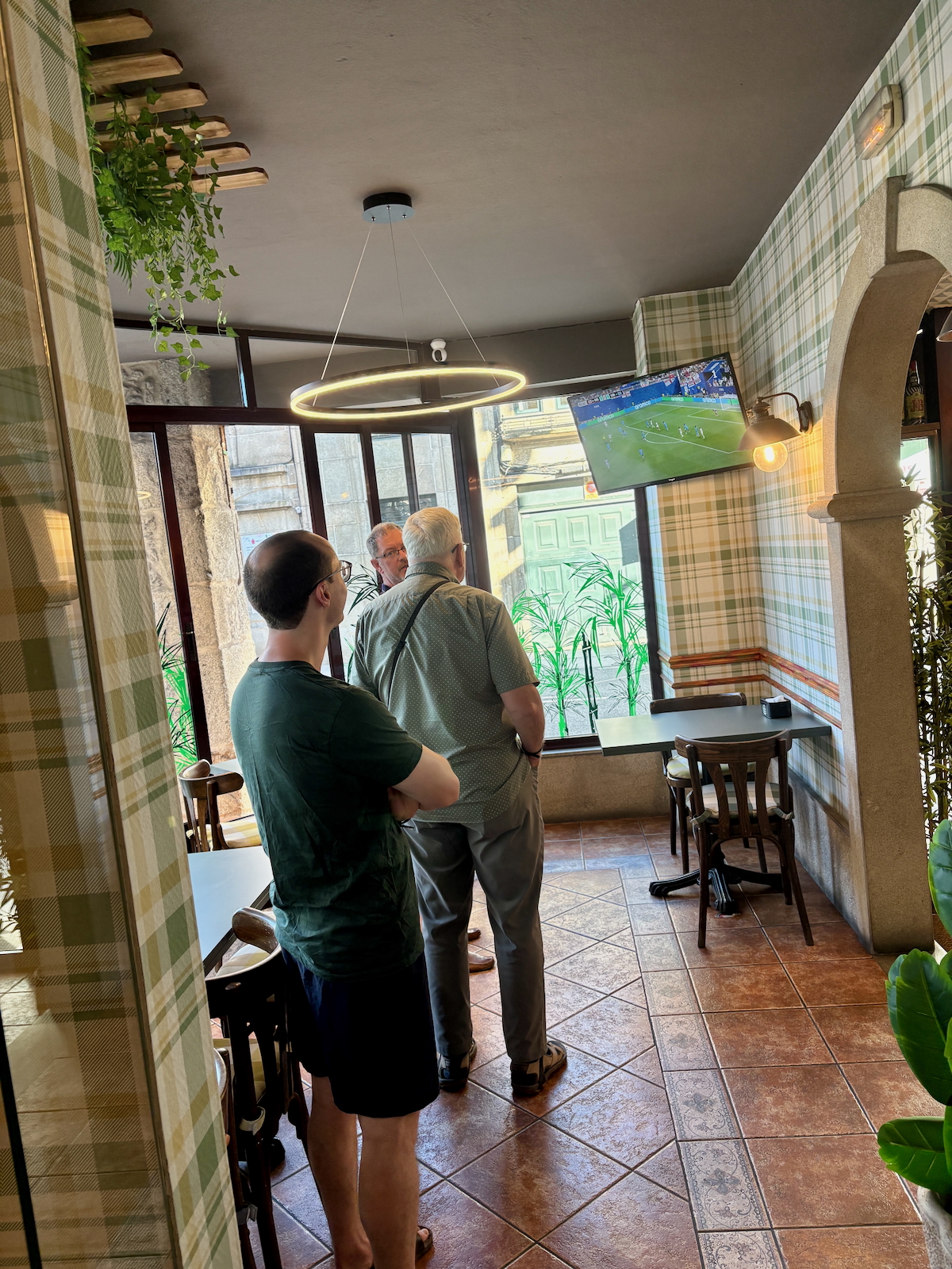 Three men stand and watch a soccer game on a wall-mounted TV in a cozy, plaid-walled cafe with warm lighting and plants nearby.