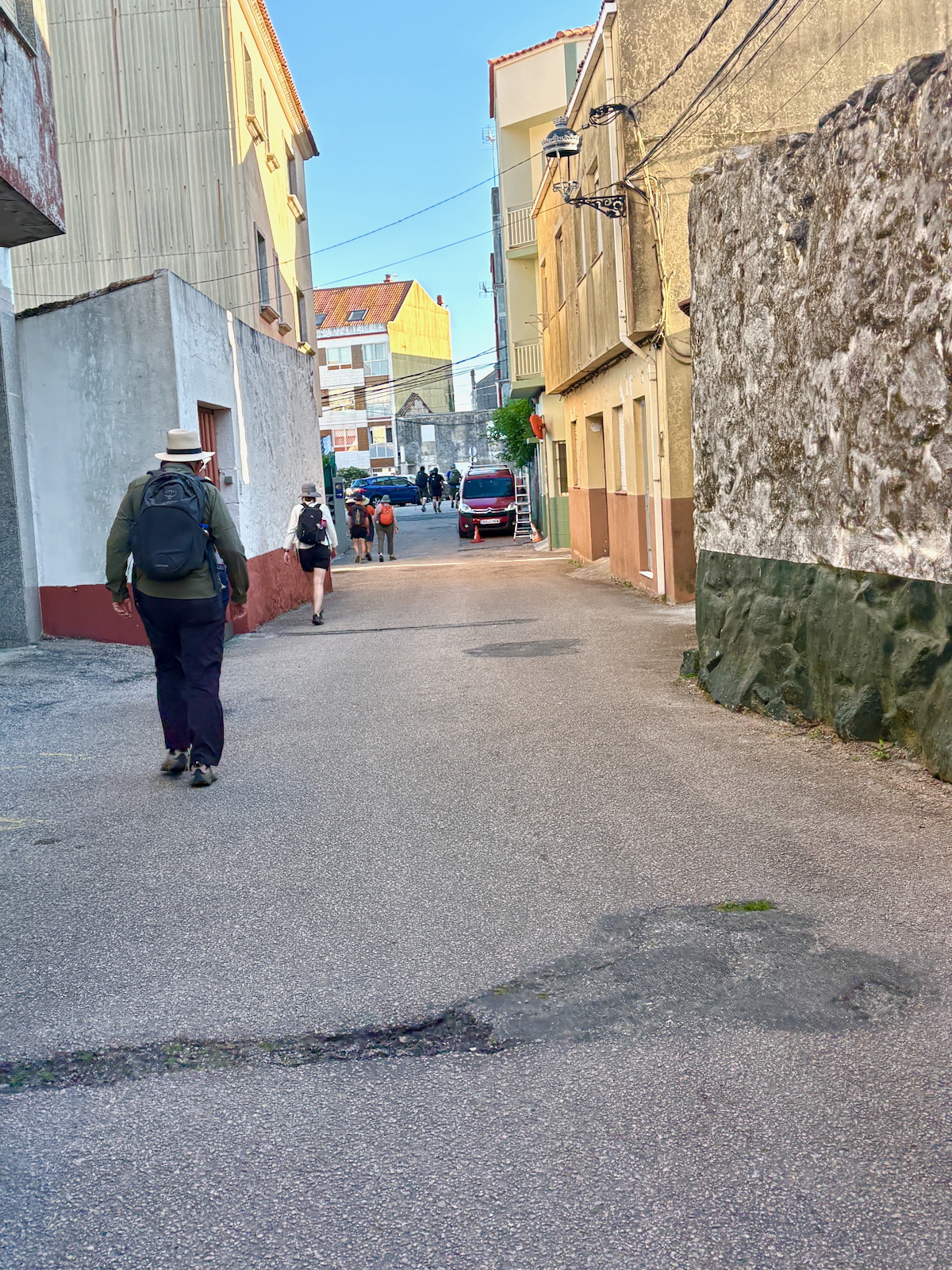 Hikers with backpacks walk down a narrow urban street lined with buildings and a stone wall on the right under a clear sky
