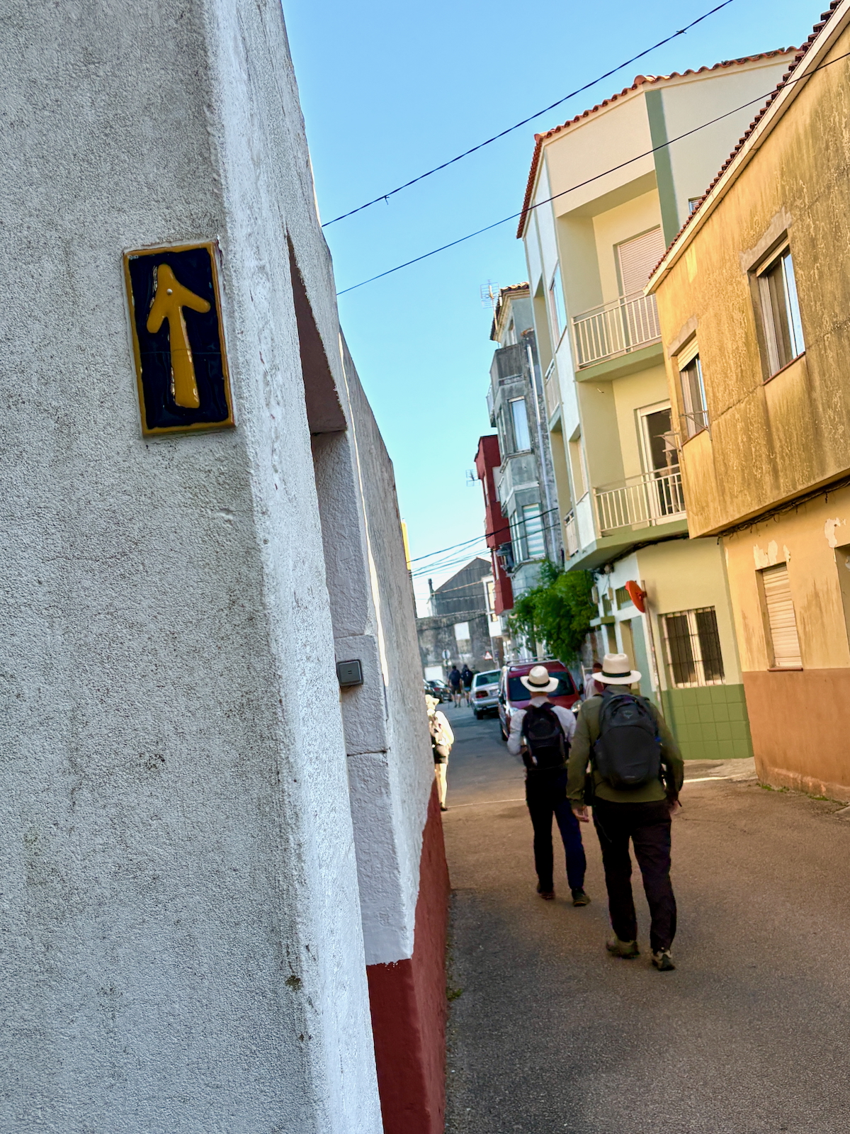 Two hikers with backpacks walk down a narrow sunlit street between white and colorful buildings; a yellow up-arrow sign is on the left wall.