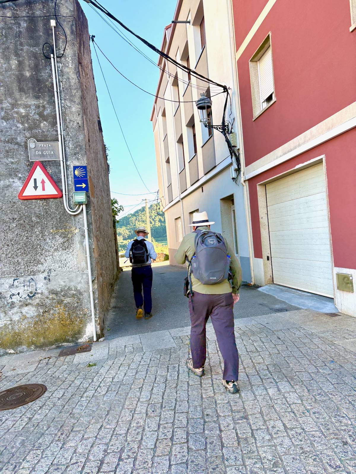 Two hikers with backpacks walk down a narrow cobblestone alley between gray wall and pastel buildings, with signs on the left wall.