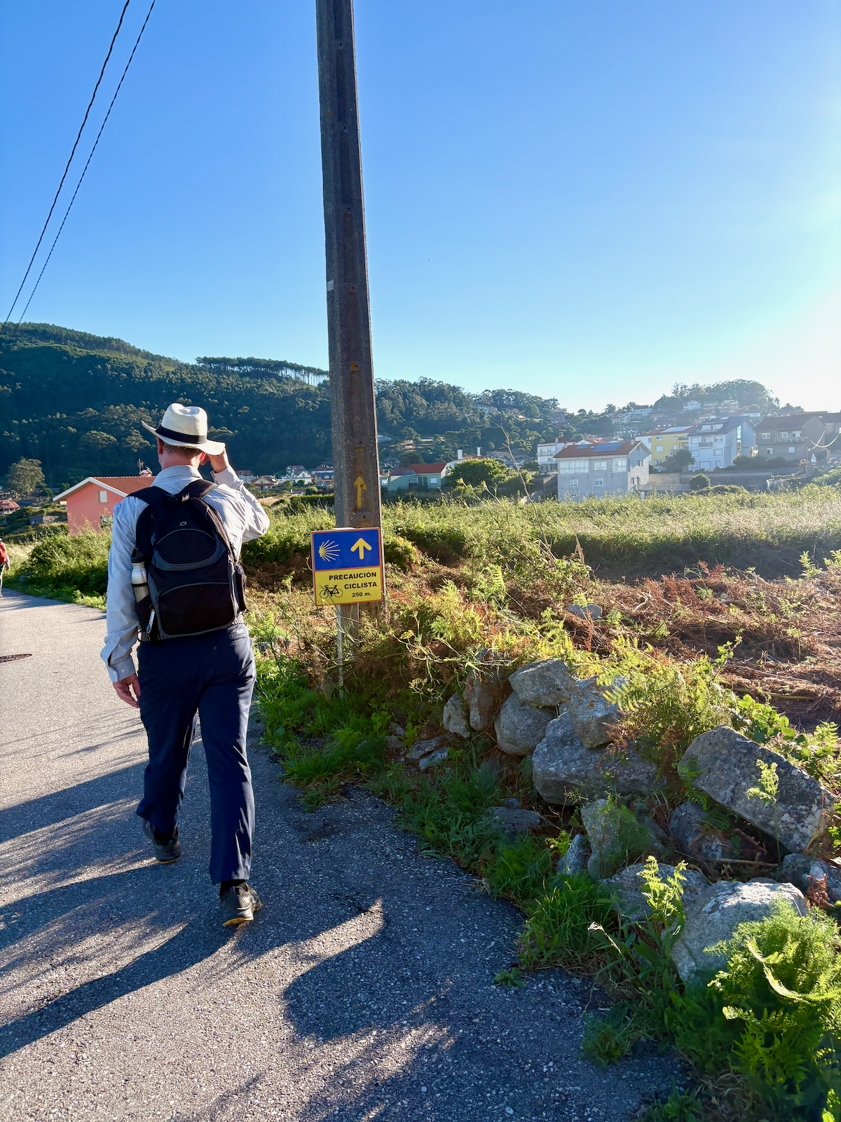 Hiker with backpack and hat walking along a rural road, yellow cyclist caution sign on a pole nearby.