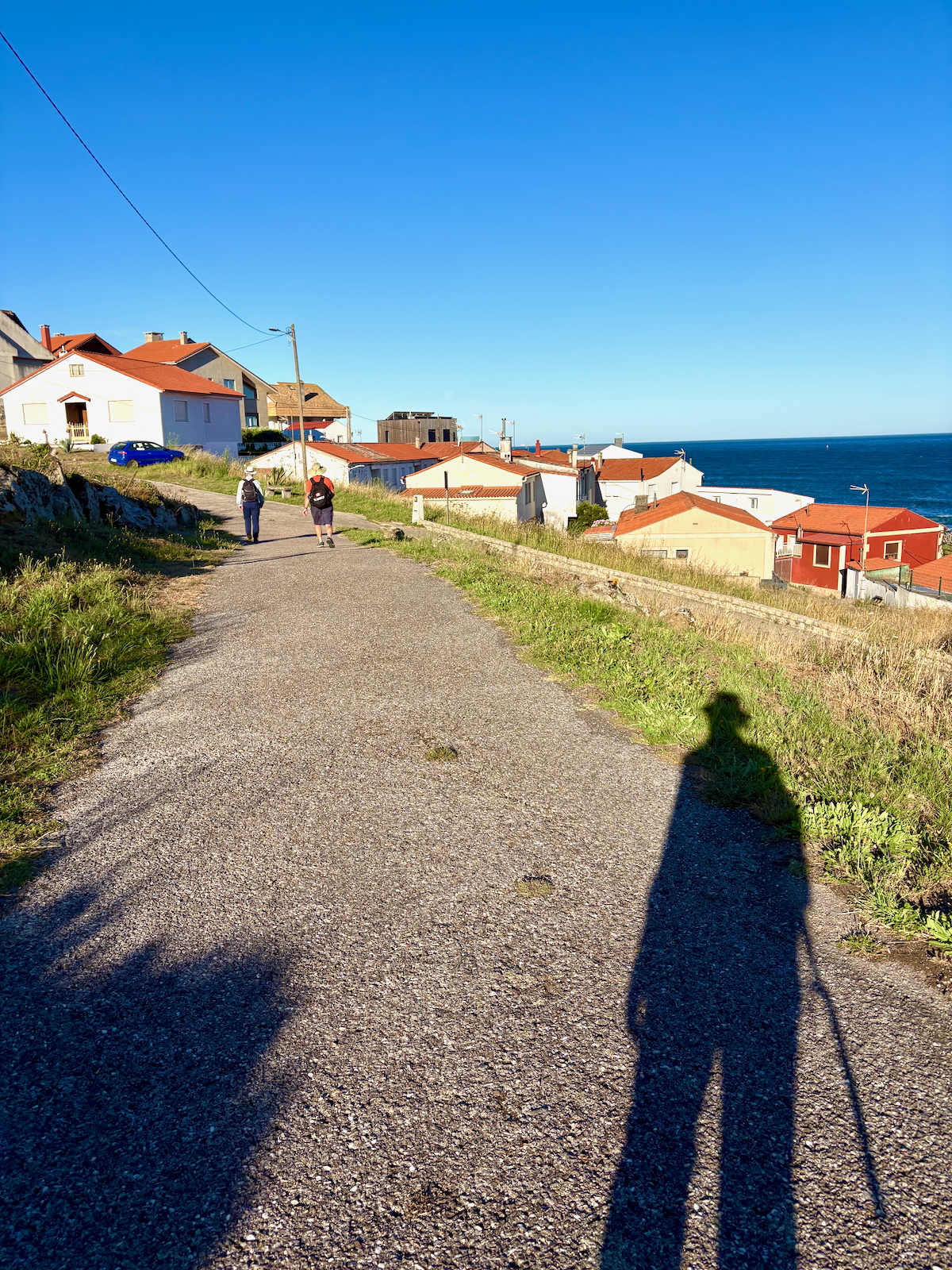 Sunlit coastal path with red-roofed houses and two hikers walking toward the sea on a clear day, blue ocean to the right.