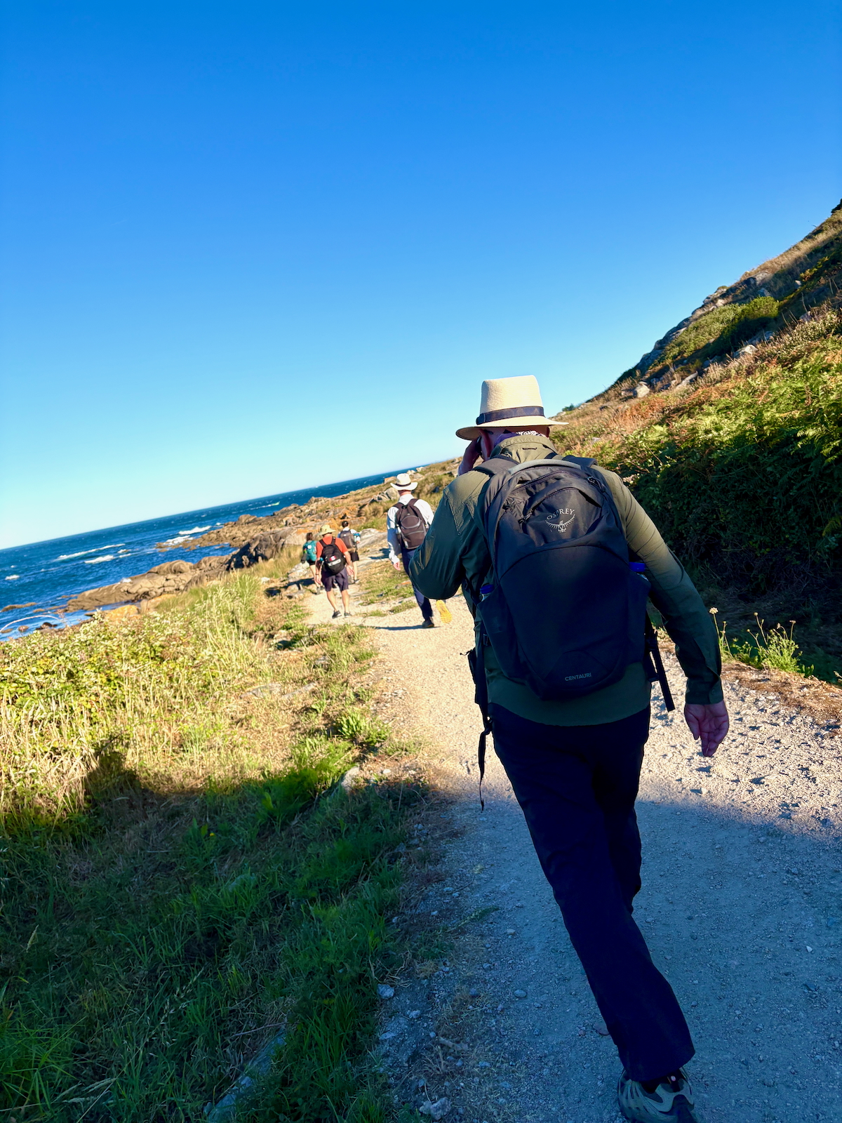 Hikers with backpacks walking along a coastal dirt path beside the blue ocean, hillside on the right under a clear sky.