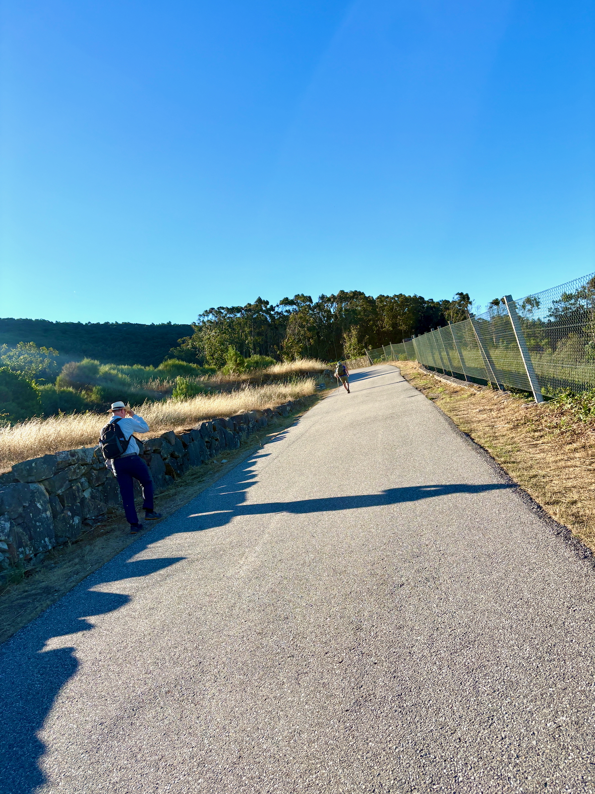 Two hikers on a sunny paved trail; one with a backpack taking a photo near a stone wall, with a chain-link fence on the right.