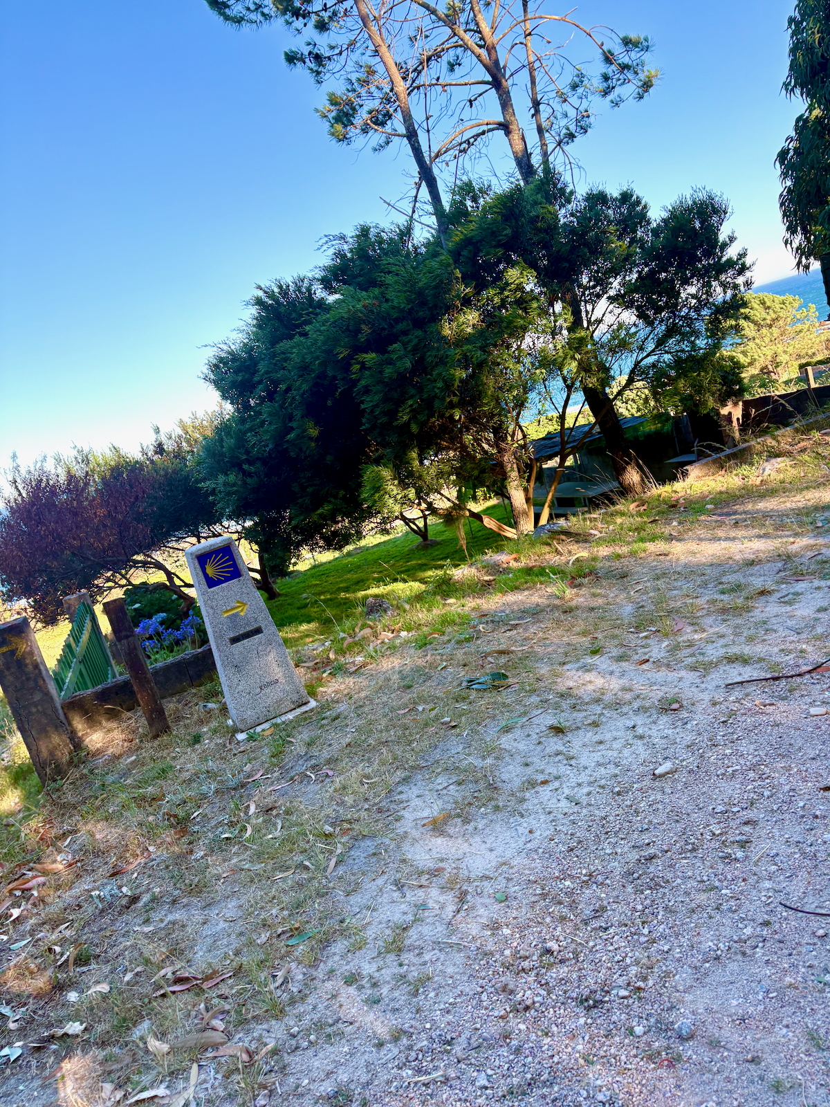 Stone trail marker with a yellow arrow and shell symbol, leaning beside a dirt path under a clear blue sky and trees.