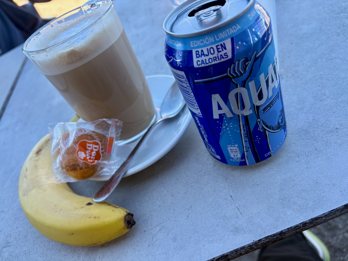 Blue can of low-calorie soda 'Bajo en calorías' beside a mug of coffee, a banana, a wrapped biscuit, and a spoon on a gray table.