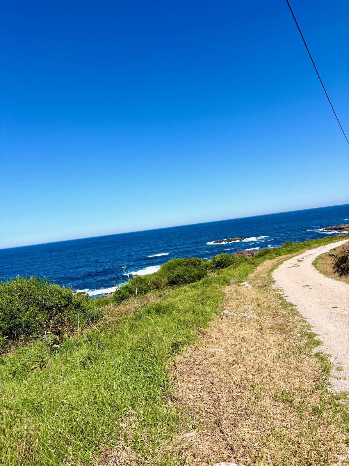 Gravel coastal path winding along a grassy cliff edge, with the blue ocean to the left and a clear sky above.