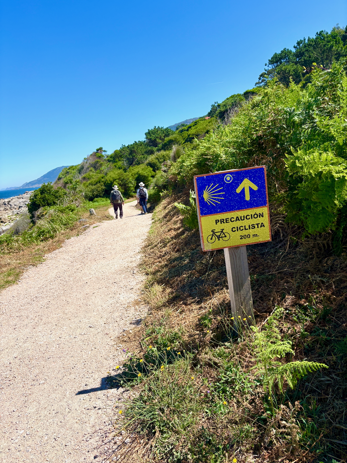 Two hikers walking along a coastal dirt trail with green hills on the right and blue sky above; a warning sign about cyclists nearby reads 'Precaución Ciclista 200 m'.