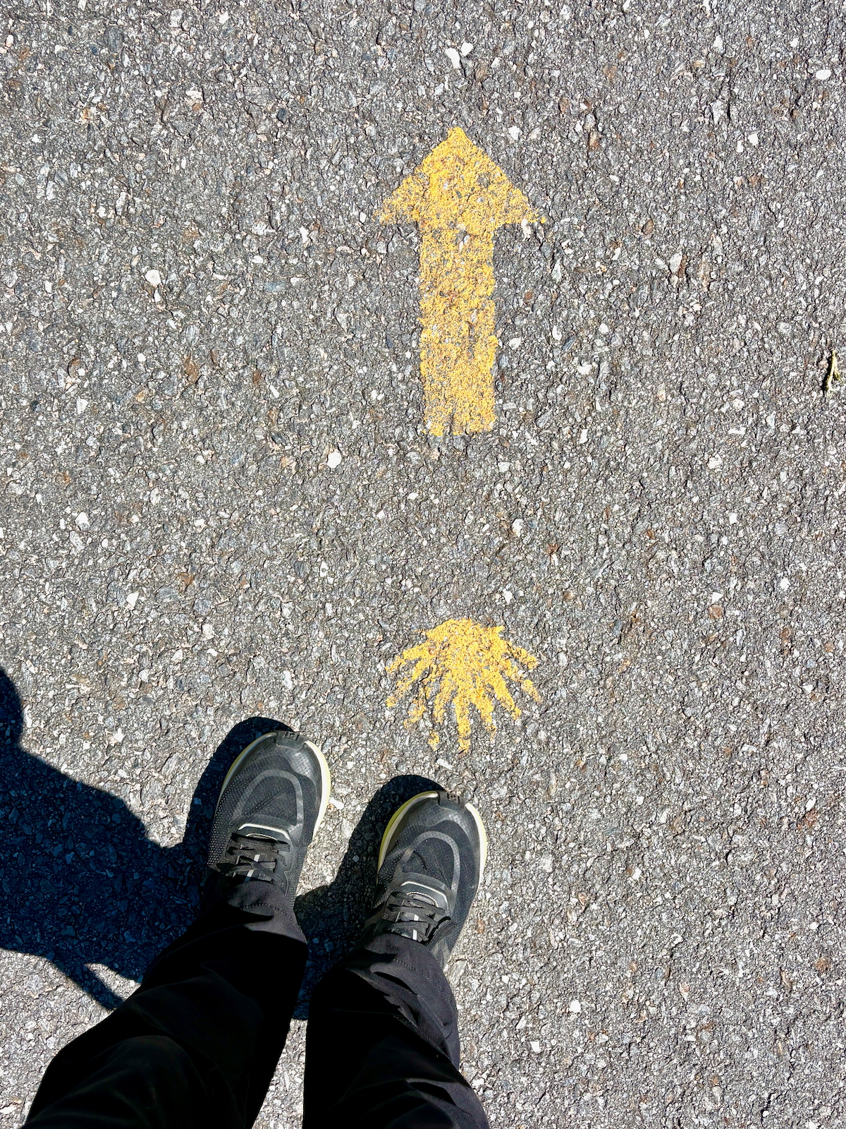 Yellow up-arrow painted on rough asphalt, with a smaller yellow palm/hand-shaped symbol nearby; photo shows a person wearing sneakers standing at the bottom edge, viewed from above.