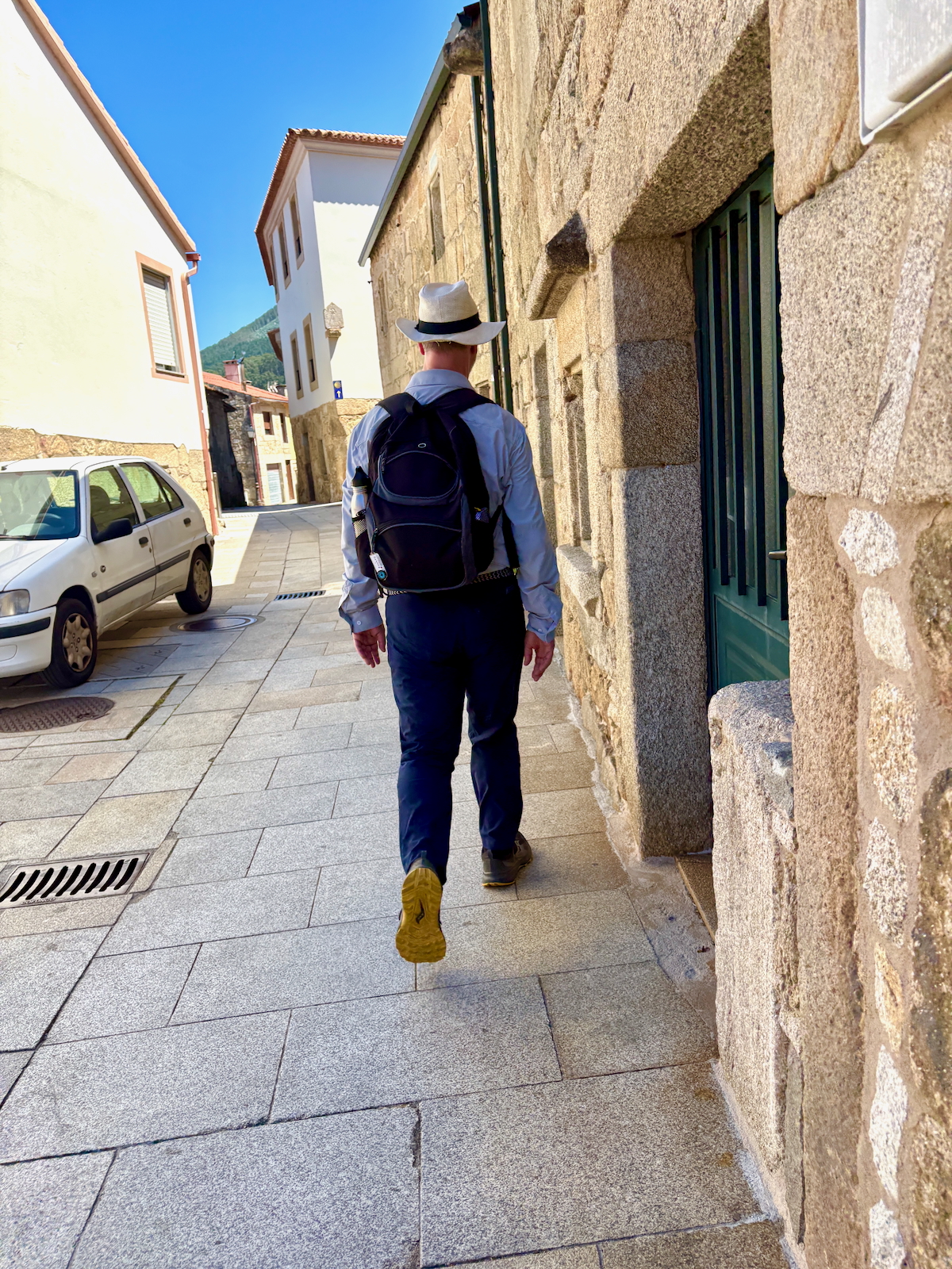 Man walking away down a narrow stone street with a backpack and wide-brim hat, white shirt, blue pants, yellow shoes, beside stone buildings.