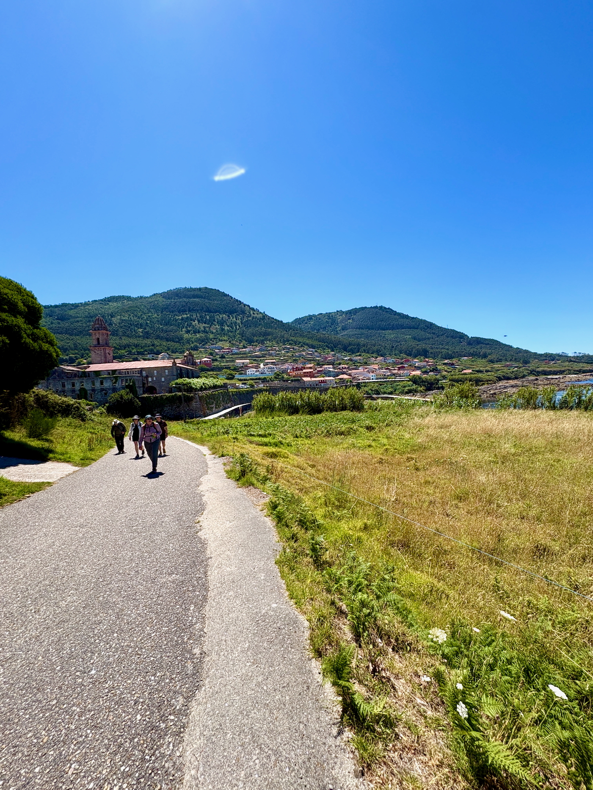 Four hikers walk along a winding country road toward a hillside village with a church tower under a bright blue sky.