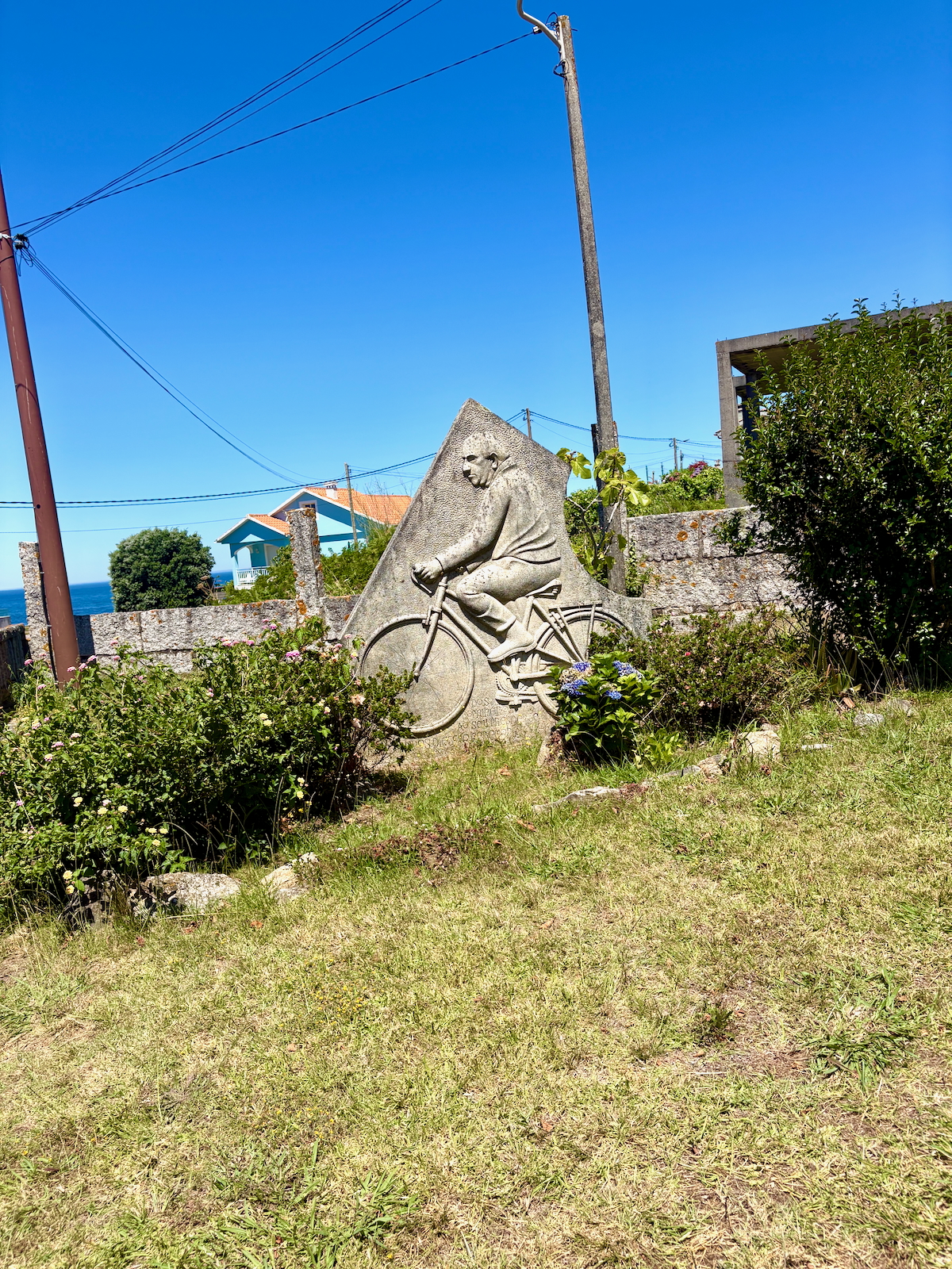 Stone sculpture of a man riding a bicycle carved into a triangular rock, surrounded by garden plants in a sunny coastal village setting.