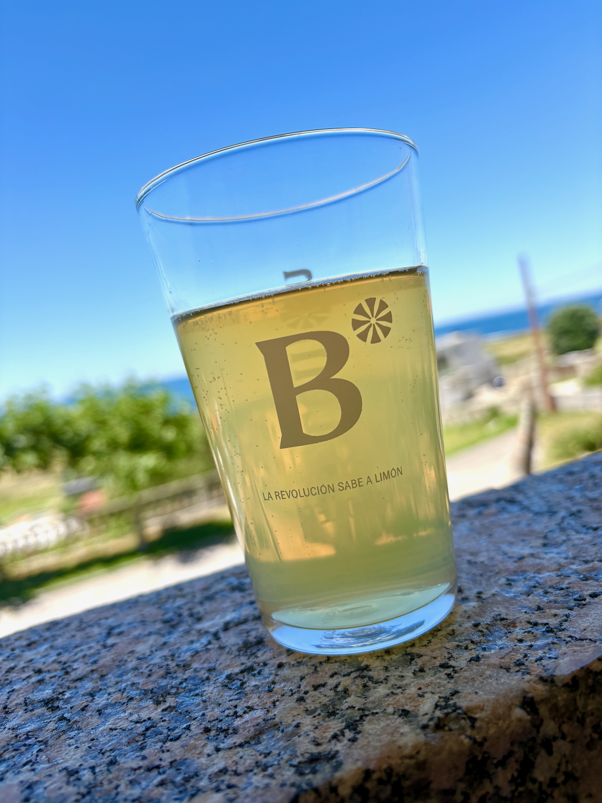 Pale lemonade in a tilted glass on a granite ledge with a blue sky, featuring a large 'B' logo and the slogan 'La revolución sabe a limón'.
