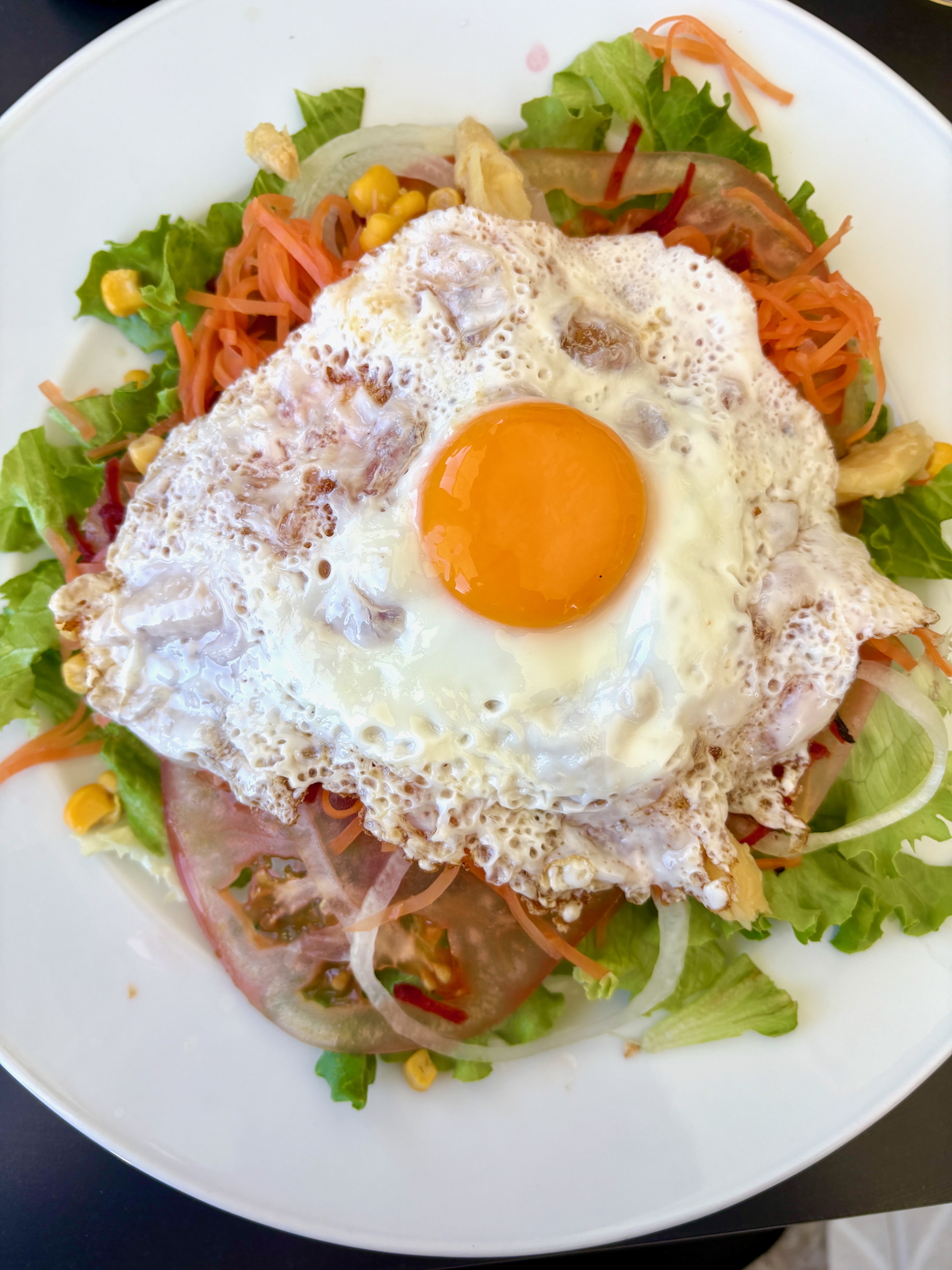 Sunny-side-up fried egg atop a mixed green salad with carrots, corn, tomatoes and onions on a white plate.