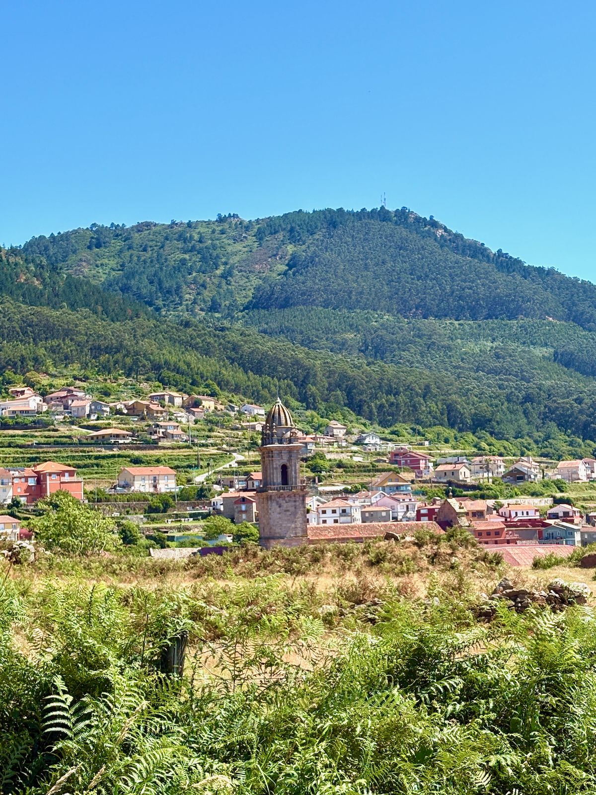 Small village at the foot of green hills with a stone church tower in the foreground and a clear blue sky.